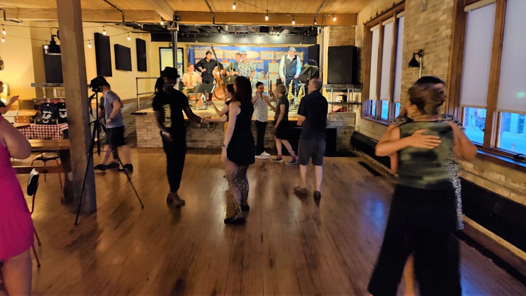 Several dancers are mid-move in front of a stage with a band playing